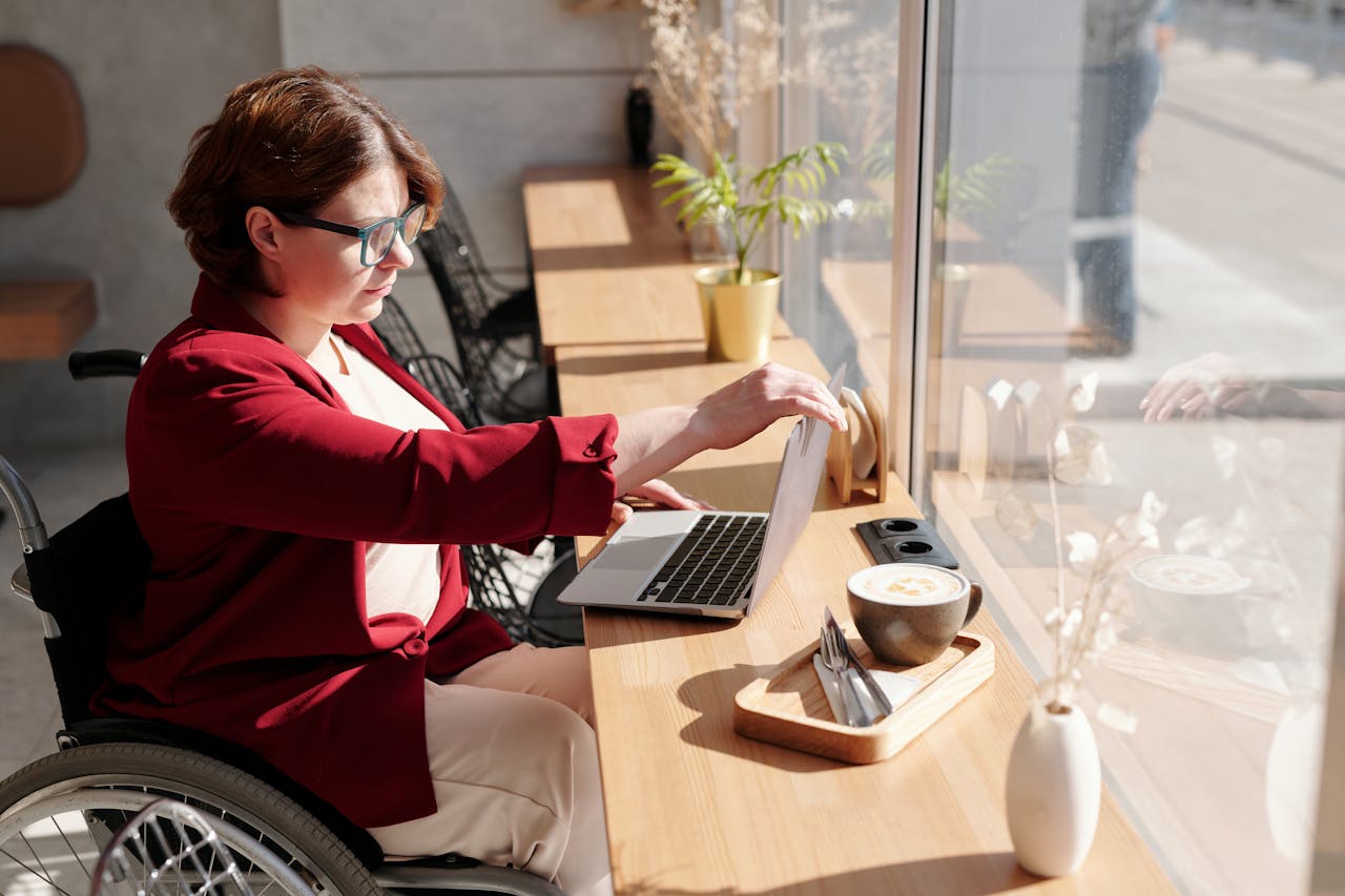 Woman in wheelchair working on a laptop in a bright café, showcasing remote work lifestyle.